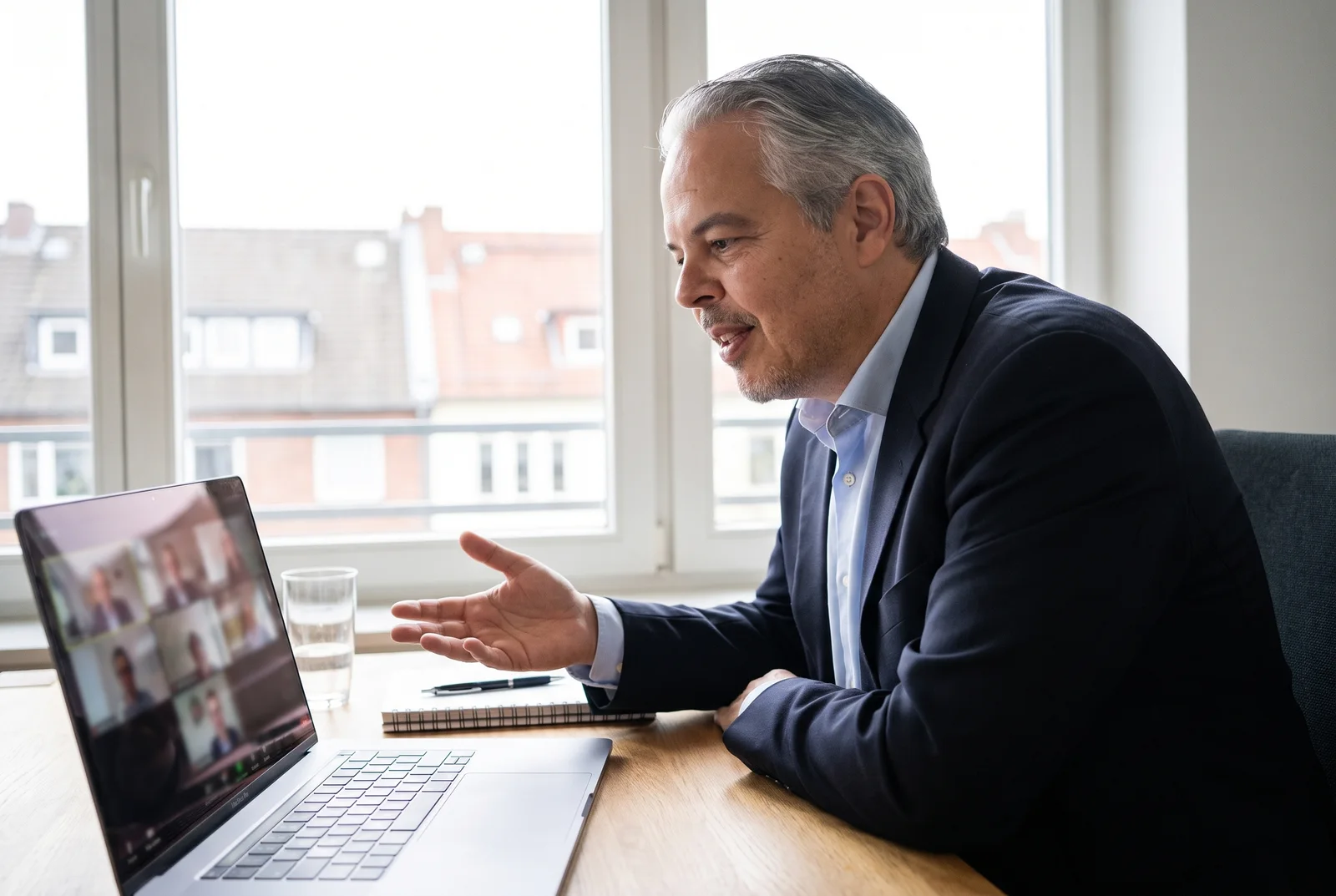 Alvaro Moreno sitzt am Schreibtisch vor Laptop und Bildschirm in einer Videokonferenz mit Kollegen und lacht, Tageslicht, kein Papier auf dem Tisch