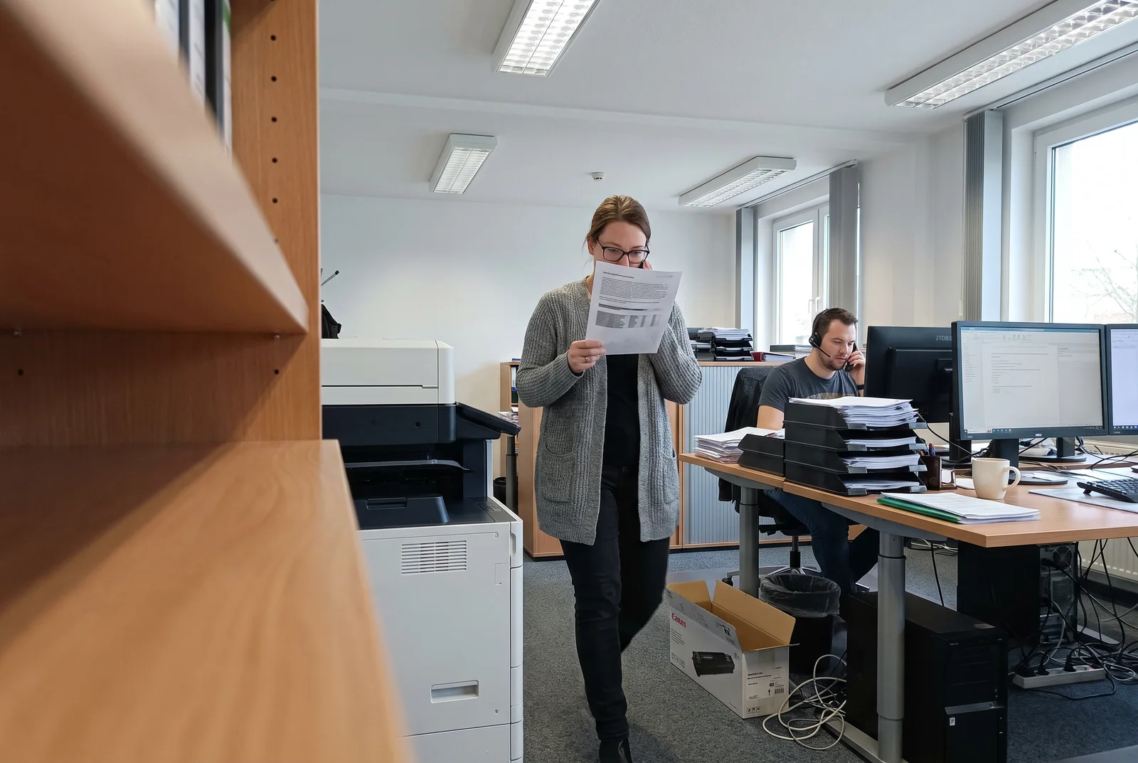 Eine Mitarbeiterin nimmt am Drucker eine ausgedruckte E-Mail aus dem Fach, im Hintergrund Schreibtische mit Bildschirmen