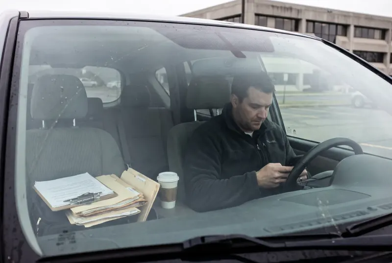 Ein Betriebsinhaber sitzt im Auto auf dem Firmenparkplatz und sucht auf dem Smartphone seine eigene Leistung, auf dem Beifahrersitz liegen Unterlagen und ein Klemmbrett