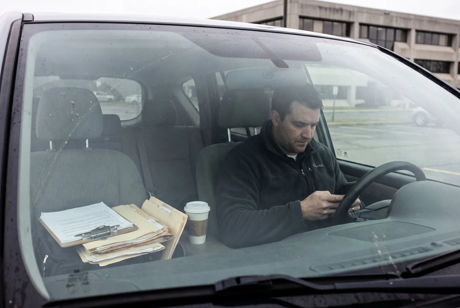 Ein Betriebsinhaber sitzt im Auto auf dem Firmenparkplatz und sucht auf dem Smartphone seine eigene Leistung, auf dem Beifahrersitz liegen Unterlagen und ein Klemmbrett