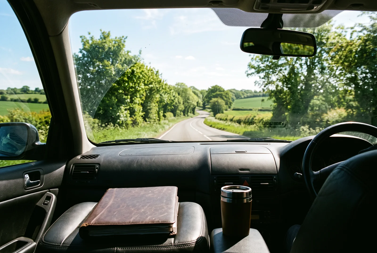 Blick aus einem Auto auf eine sonnige Landstraße am Vormittag, auf dem Beifahrersitz eine Mappe und ein Becher