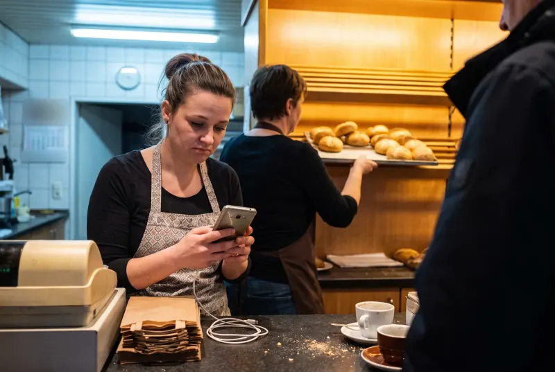 Verkäuferin in einer Bäckerei sieht stirnrunzelnd auf ein älteres Firmenhandy, hinten legt eine Kollegin Brötchen ins Regal