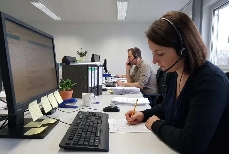 Eine Mitarbeiterin im Kundendienst mit Headset macht während eines Telefonats einen Strich auf einer handgeschriebenen Liste neben der Tastatur