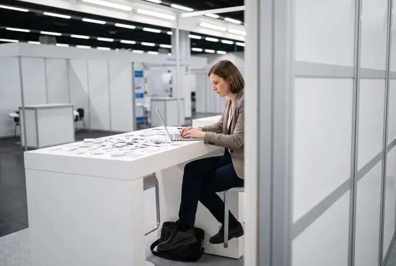 Eine Frau sitzt am Abend allein am Messestand und trägt hunderte vor ihr ausgebreitete Visitenkarten in den Laptop ein